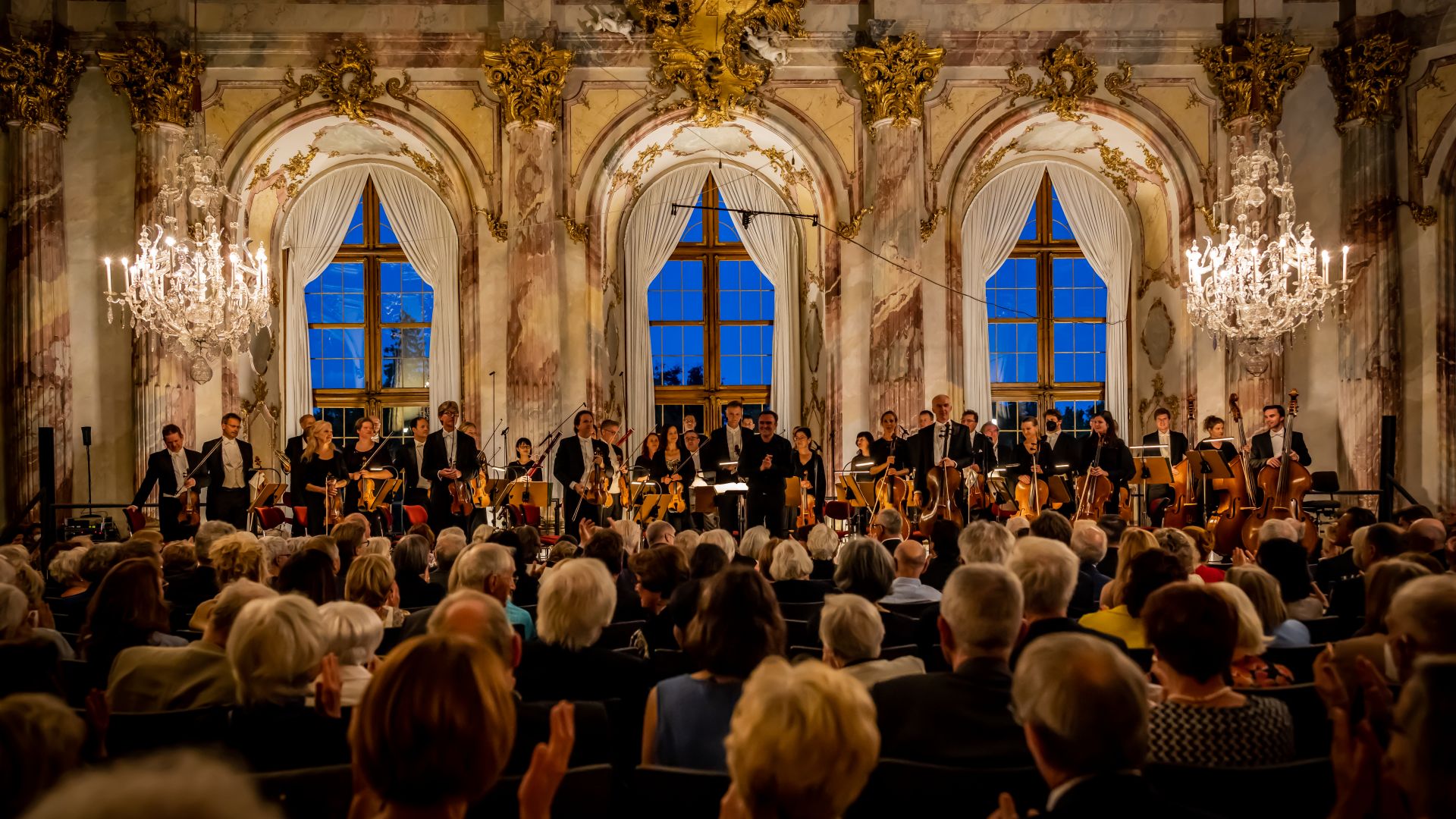 The image shows the magnificent setting of the Würzburg Residence during the Mozart Festival. In a grand Baroque hall, an orchestra performs classical music for an enthusiastic audience.