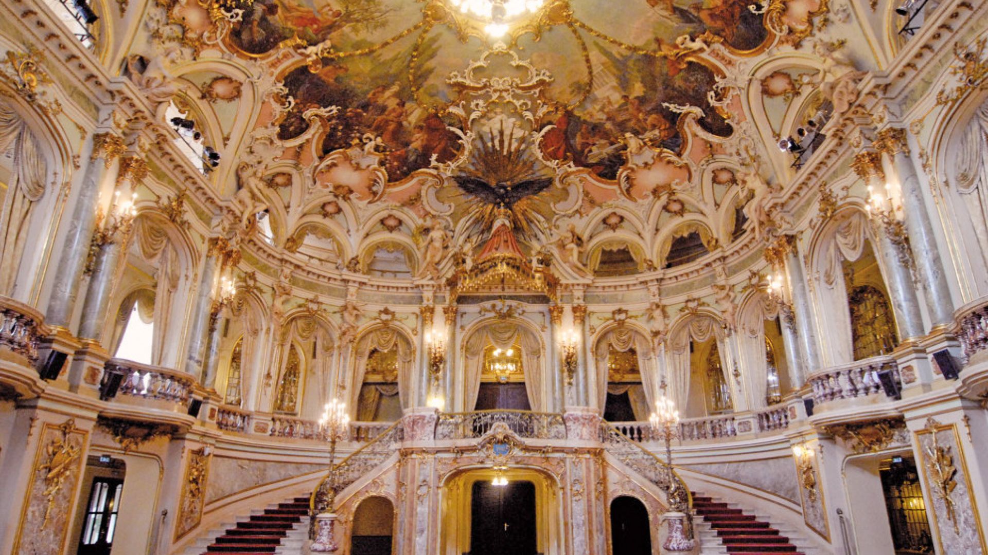 The image shows the magnificent interior of the Hessian State Theatre with elaborately decorated walls, a grand staircase, and a beautifully painted Baroque-style ceiling.