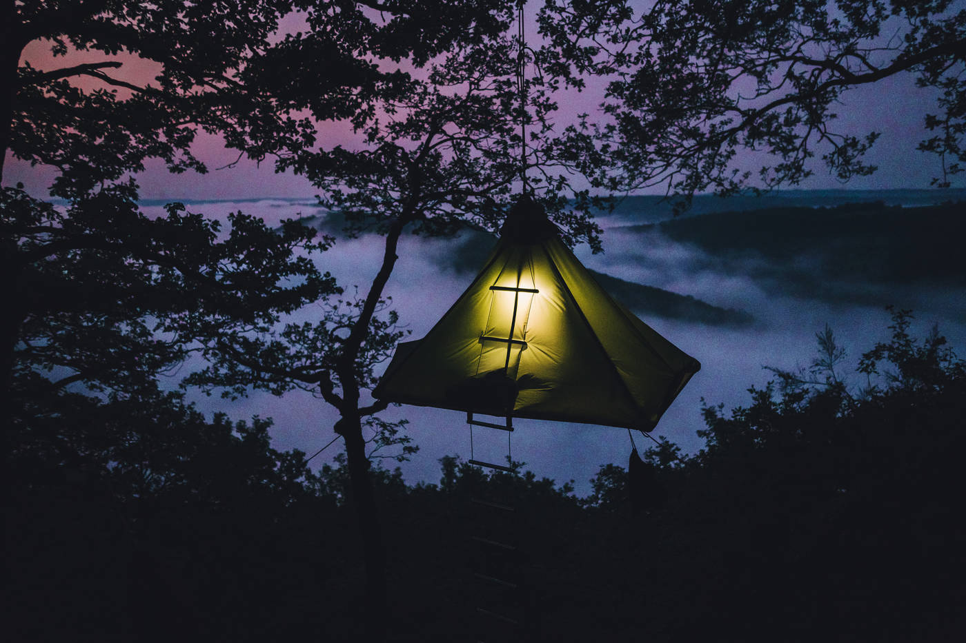 Una tienda iluminada colgada entre los árboles por la noche, por encima de la niebla en Sarre.