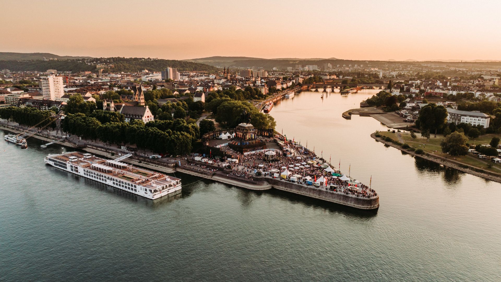 Luftaufnahme des Electronic Wine Festivals in Koblenz bei Sonnenuntergang, mit Menschen am Deutschen Eck und Schiffen auf dem Rhein.