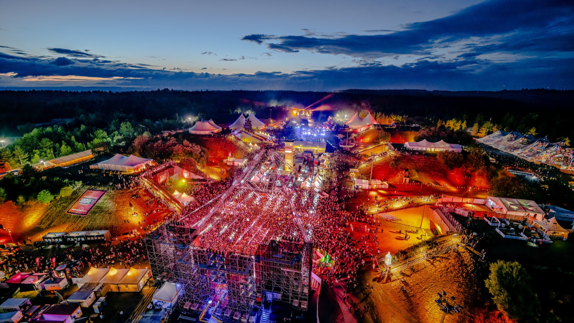 Luftaufnahme des NatureOne Festivals bei Nacht mit beleuchteten Bühnen und einer großen Menschenmenge.