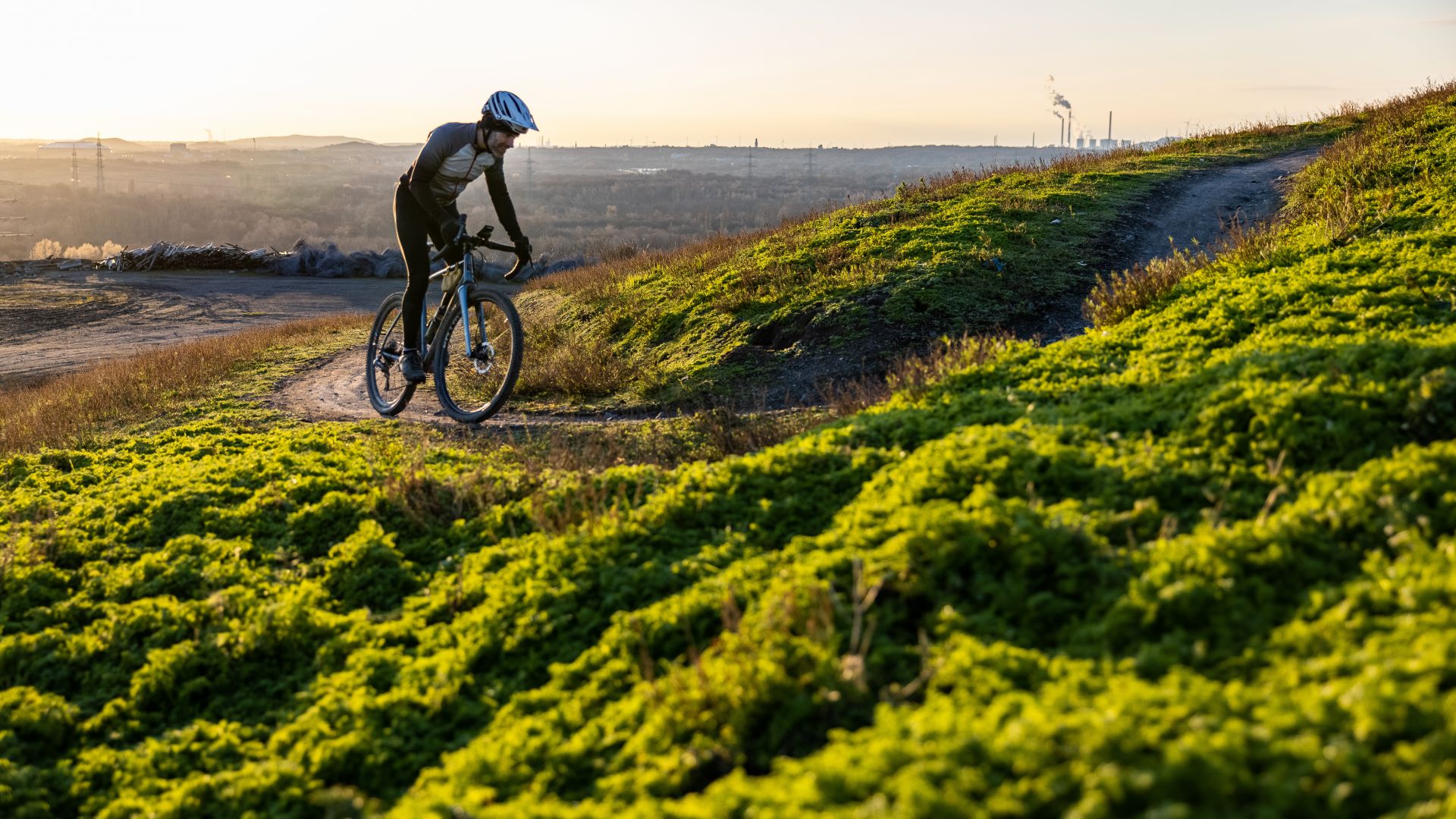 Un gravel biker recorre un camino de grava en el montículo de Hoheward en Herten, rodeado de vegetación verde y con vistas al paisaje distante al atardecer.