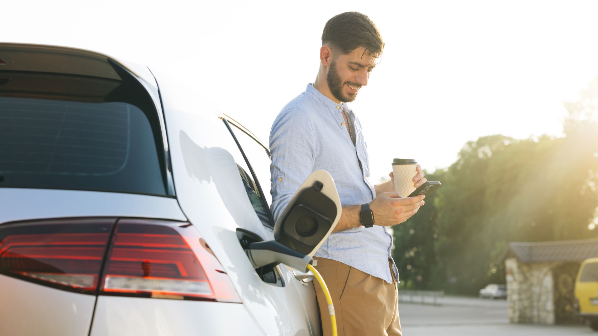 Un homme avec un smartphone et un café se tient à une borne de recharge avec une voiture électrique