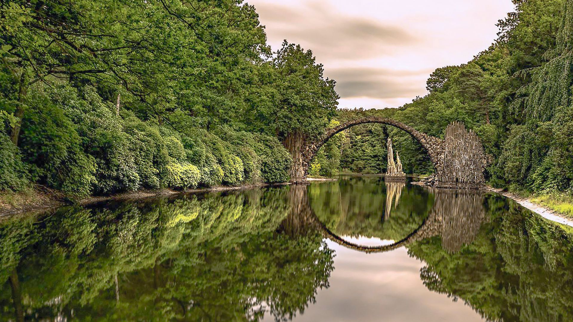 Rakotz Bridge in Kromlau reflecting over a calm lake, surrounded by dense forest.